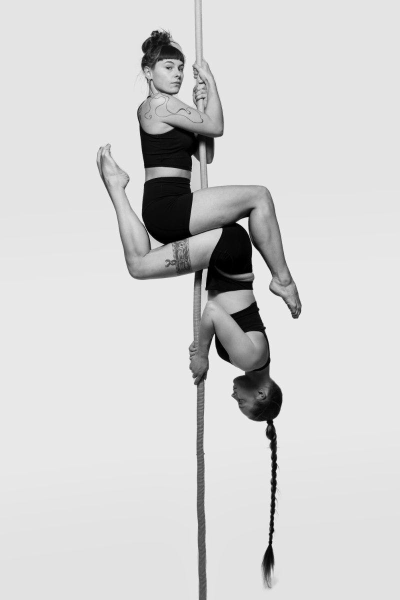Melinda and another performer hanging from a rope. Both performers have a seated position and are in such a way that the picture could be upside down. One is sitting on the other or possibly vice-versa.