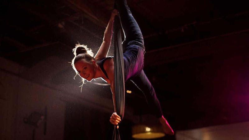 Melinda upside-down in a set of aerial silks. She is being held up by her left foot which is lifted up behind her and hooked to the silks.