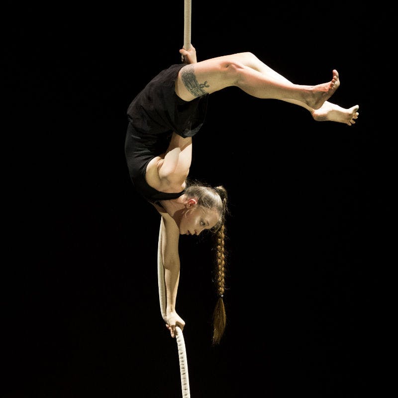 Melinda performing a trick called the scorpion on a rope. It is as if she is performing a handstand on the rope with her legs bent backwards over her head to keep her stable. She is wearing a black dress and her hair is flowing below her.